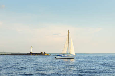 White sloop rigged yacht sailing in the Baltic sea at sunset. Clear sky after the storm, soft sunlight. Lighthouse. Transportation, travel, cruise, sport, recreation, leisure activity, racing, regattaの写真素材