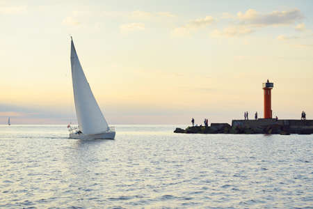 White sloop rigged yacht sailing in the Baltic sea. Lighthouse, walking people. Clear golden sunset sky after the storm. Transportation, travel, cruise, sport, recreation, leisure activity, regattaの写真素材