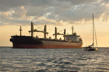 Large cargo ship (bulk carrier, 179.99 meters length) and sloop rigged yacht sailing in the Baltic sea at sunset. Freight transportation, global communications, economy, cruise, recreation. sportの写真素材