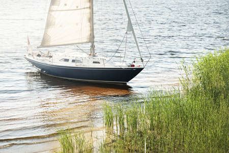 Blue sloop rigged yacht anchored to the sandy shore (beach overgrown with grass). Sailing in Sweden,...