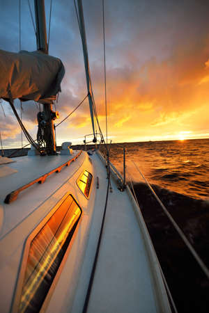 White yacht sailing in an open sea at sunset. A view from the deck to the bow, mast, sails. Epic cloudscape. Dramatic sky with glowing golden clouds after the storm. Racing, sport, leisure activityの写真素材