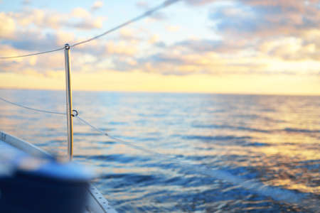 White sloop rigged yacht sailing in an open sea at sunset. Boat side railing close-up. Dramatic sky, glowing clouds. Transportation, travel, cruise, sport, recreation, leisure activity, regattaの写真素材
