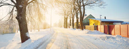 Snow-covered country road through the village of wooden traditional houses. Tall trees in hoarfrost, warm sunlight. Clear winter day. Latvia