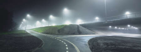 An empty illuminated bicycle road with a sharp turn in a fog at night. Lanterns close-up. Bridge in...