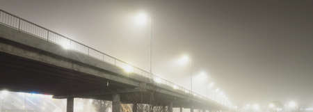 Low angle view of the illuminated empty road bridge in a fog at night. Lanterns close-up. Panoramic imageの写真素材