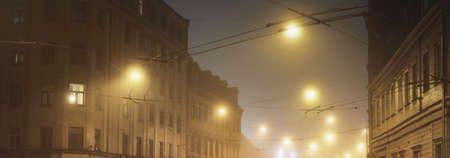 An empty illuminated asphalt road through the old historical buildings and houses in a fog at night....
