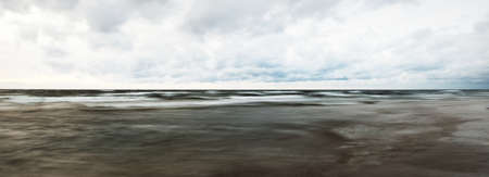 Baltic sea shore after the storm. Dramatic sunset sky, dark glowing clouds, golden sunlight. Waves, splashing water. Picturesque scenery, seascape, cloudscape, nature. Panoramic view, long exposureの写真素材