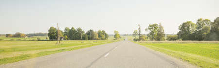 Country asphalt road (highway) through the field and forest on a clear day. Warm midday sunlight, clear sky. A view from the car. Travel destinations, vacations, freedom, driving, transportationの写真素材