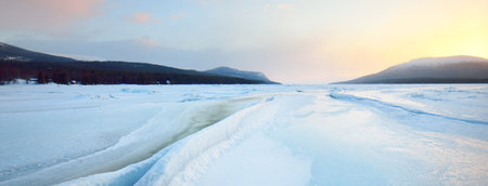 Ice pressure ridge on a frozen lake shore at sunset. Mountain peaks in the background. Colorful cloudscape. Picturesque winter scenery. Ecology, environment, climate change, global warming conceptsの写真素材