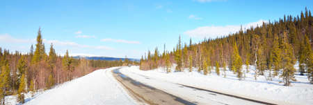 Empty old asphalt road (highway) through the pine forest. Snow-covered mountains. Winter landscape. Kola Peninsula, Polar Circle, Russia. Expedition, work, route, logistics, remote places, off-roadの写真素材