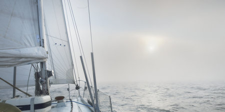White yacht sailing in an open Baltic sea in a thick fog at sunrise. Germany. Close-up view from the deck to the bow and sails. Morning sun and glowing clouds. Epic seascape. Sport and recreation themeの写真素材