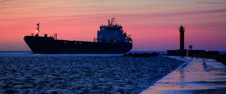 Large cargo ship sailing near the lighthouse at sunset. Colorful cloudscape. Riga bay, Baltic sea, Latvia. International communications, logistics themeの写真素材