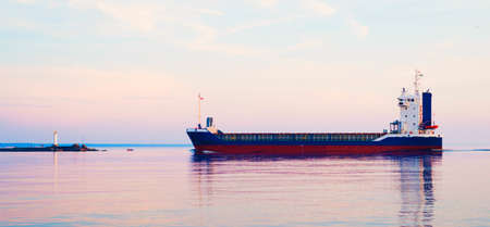 Large cargo ship sailing in the Baltic sea at sunset. Soft golden sunlight. Concept seascape. Panoramic view from the sailing boat. Freight transportation, nautical vessel, logisticsの写真素材