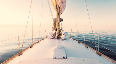 White yacht sailing in a still water at sunset. A view from the deck to the bow, mast, sails. Stunning cloudscape. Reflections on water. Baltic sea. Lifestyle, cruise, regatta, sport, recreationの写真素材