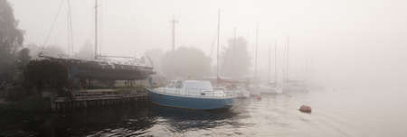 Sailing boats moored to a pier in a thick white morning fog at sunrise, close-up. Yacht club in...