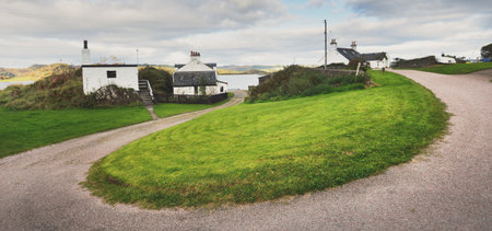 An empty country road and small traditional house on the rocky shore of Crinan canal. Hills and...