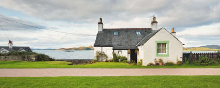 An empty country road and small traditional house on the rocky shore of Crinan canal. Hills and forest in the background. Dramatic cloudscape. Travel destinations theme. Scotland, UKの写真素材