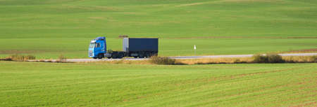 Modern semi-trailer truck riding through green plowed agricultural field with tractor tracks. Forest...