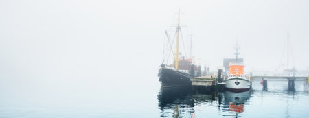 Small ships, yachts, sailing and fishing boats moored to a pier, in a thick white morning fog, close-up. Kiel, Germany. Transportation, logistics, industry, cityscape. Panoramic imageの写真素材