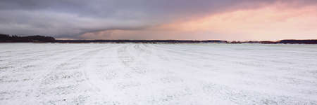 Snow-covered plowed country agricultural fields with tractor tracks. Dramatic sunset sky, colorful clouds. Winter landscape. Latvia. Warm winter, climate change, global warmingの写真素材