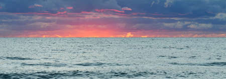 Baltic sea shore at sunset. Soft sunlight, dramatic sky, glowing pink and golden clouds. Waves and water splashes. Idyllic seascape. Weather, climate change, nature. Panoramic view, copy spaceの写真素材