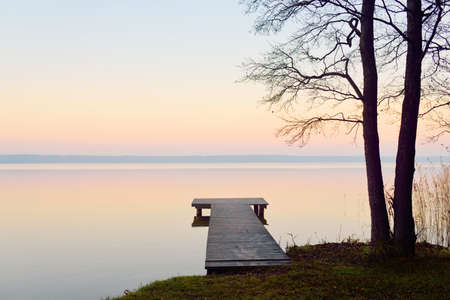 Forest lake (river) at sunrise. Wooden pier (boardwalk), mighty trees. Soft sunlight, mist, reflections on water. Idyllic autumn landscape. Pure nature, ecotourism, hiking, exploringの写真素材