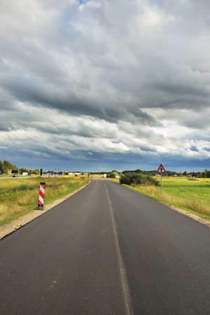 Country highway (asphalt road) through the fields and forest. View from the car. Dramatic storm clouds. Travel destinations, route, vacations, logistics, remote placesの写真素材