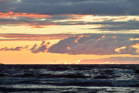 Panoramic view from the Baltic sea shore at sunset. Dramatic storm sky, glowing clouds, golden sunlight. Waves, splashing water. Nature, environment, fickle weatherの写真素材