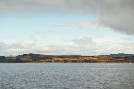 Dramatic sky above the rocky shores, hills and valleys of Rothesay, Scotland. Panoramic view from a sailing boat. Travel destinations, tourism, hiking, landmark, ecology, environment, nature themesの写真素材