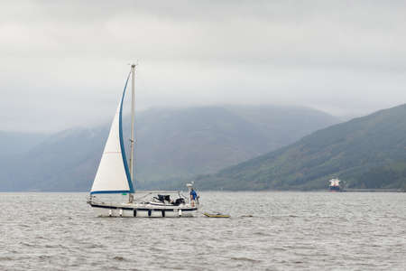Yacht sailing near the rocky lake shore. Rothesay, Scotland, UK. Nautical vessel, transportation,...
