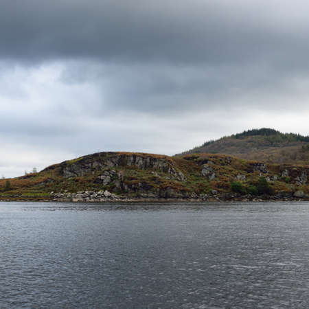 Panoramic view of the rocky shores of Kyles of Bute from the water. Hills and mountains in the background. Dark storm sky. Bute island, Firth of Clyde, Scotland, UKの写真素材