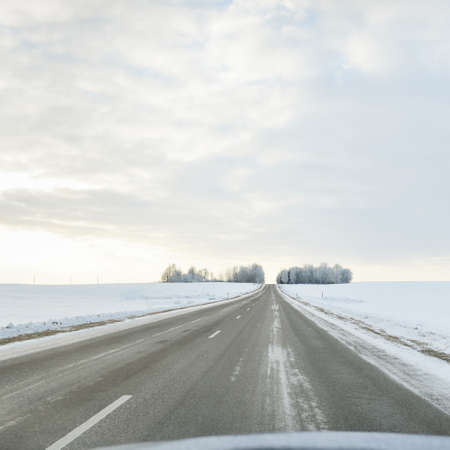 Empty highway (asphalt road) through the snow-covered forest and fields, rural area. Snow drifts. Europe. Nature, christmas vacations, remote places, winter tires, dangerous driving conceptの写真素材