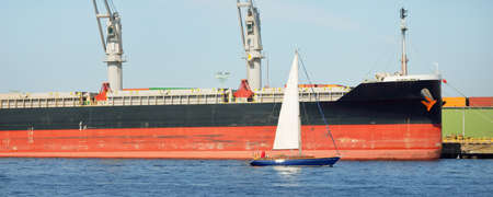 Sloop rigged yacht sailing in the Baltic sea. Cargo ship loading in port terminal in the background....