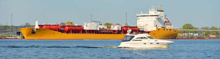 White cutter sailing in the Baltic sea. Tanker ship loading in port terminal in the background. Transportation, travel, cruise, sport, recreation, leisure activity, business, economy, industry, supplyの写真素材