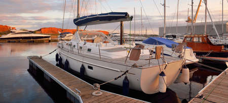 Modern sailboat with wooden teak deck moored to a pier in a yacht marina at sunset. Dramatic sky,...
