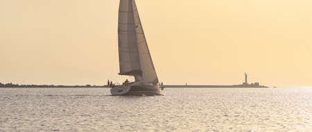 Sloop rigged yacht sailing in the Baltic sea. Lighthouse close-up. Clear golden sunset sky after the...