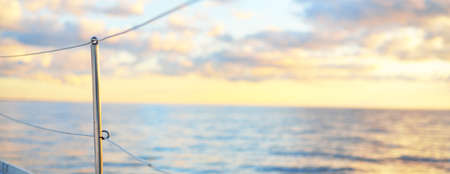 White sloop rigged yacht sailing in an open sea at sunset. Boat side railing close-up. Dramatic sky,...