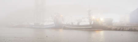 Ships, tugboats and fishing boats (trawlers) moored to a pier in a harbor. Thick white fog. Latvia, Baltic sea. Panoramic view. Service, repair, freight transportation, logistics, industry, commerceの写真素材