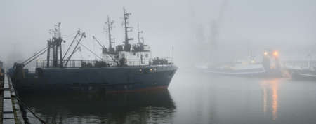 Tugboats and fishing boats (trawlers) moored to a pier in a harbor. Thick white fog. Latvia, Baltic sea. Panoramic view. Service, repair, freight transportation, logistics, industry, commerceの写真素材