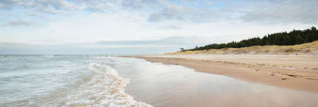 Panoramic view of the Baltic sea from a sandy shore (sand dunes). Dramatic sky with glowing clouds, waves and water splashes. Idyllic seascape. Warm winter weather, climate change, nature. Latviaの写真素材
