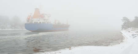 Frozen river, large cargo ship. Snow, thick fog. Concept winter urban landscape. Transportation, nautical vessel, global communications, industry, logisticsの写真素材