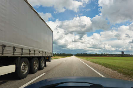 Modern semi-trailer truck riding through green plowed agricultural field and forest. Highway, freight transportation, articulated lorry, logistics, industry, delivery, remote places, route. Europeの写真素材