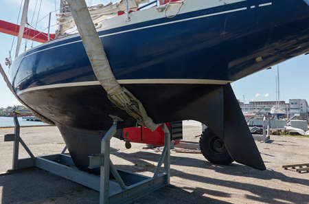 Blue sloop rigged sailboat lifted by a crane being placed back in the water after winterization. Ventspils, Latvia. Service and repair, transportation, cruise, travel, sport and recreation themesの写真素材