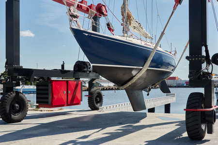 Blue sloop rigged sailboat lifted by a crane being placed back in the water after winterization. Ventspils, Latvia. Service and repair, transportation, cruise, travel, sport and recreation themesの写真素材