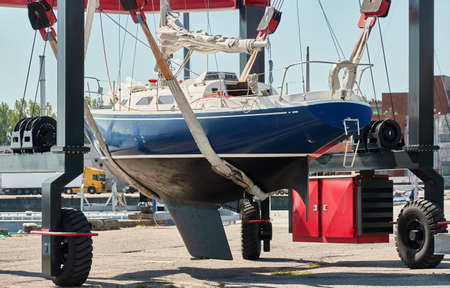 Blue sloop rigged sailboat lifted by a crane being placed back in the water after winterization. Ventspils, Latvia. Service and repair, transportation, cruise, travel, sport and recreation themesの写真素材