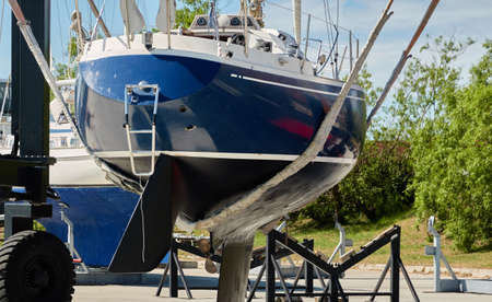Blue sloop rigged sailboat lifted by a crane being placed back in the water after winterization. Ventspils, Latvia. Service and repair, transportation, cruise, travel, sport and recreation themesの写真素材