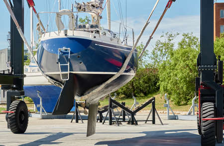 Blue sloop rigged sailboat lifted by a crane being placed back in the water after winterization. Ventspils, Latvia. Service and repair, transportation, cruise, travel, sport and recreation themesの写真素材