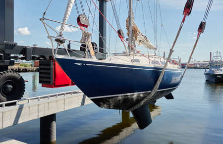 Blue sloop rigged sailboat lifted by a crane being placed back in the water after winterization. Ventspils, Latvia. Service and repair, transportation, cruise, travel, sport and recreation themesの写真素材