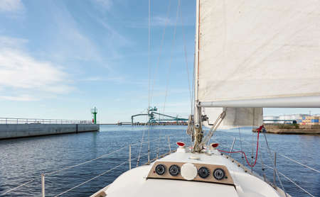 Sloop rigged yacht leaving port at sunset. Cargo terminal and ships in the background. Baltic sea....