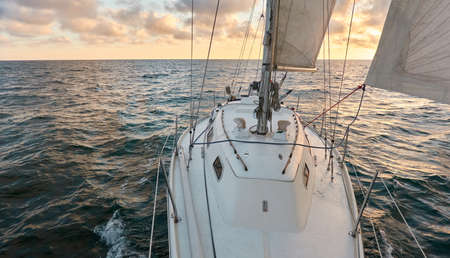Yacht sailing in an open sea at sunset. Top down view of the deck, mast and sails. Clear sky after the rain, dramatic glowing clouds, golden sunlight. Waves, water splashes. Travel, cruise, sportの写真素材